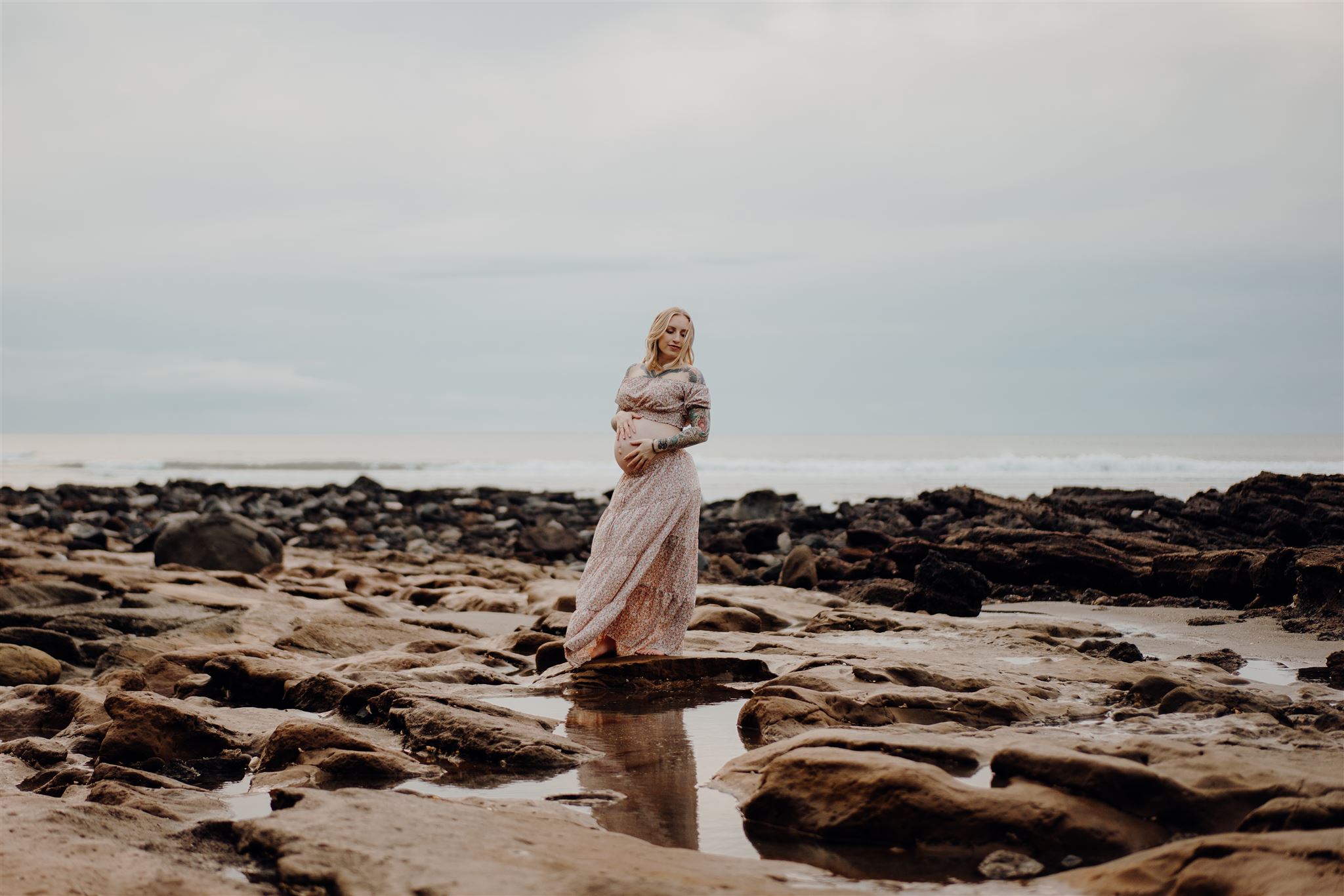 A woman wearing a maternity dress standing on the rocks at Ngarunui Beach in Raglan, Waikato being photographed by Haley Adele Photography