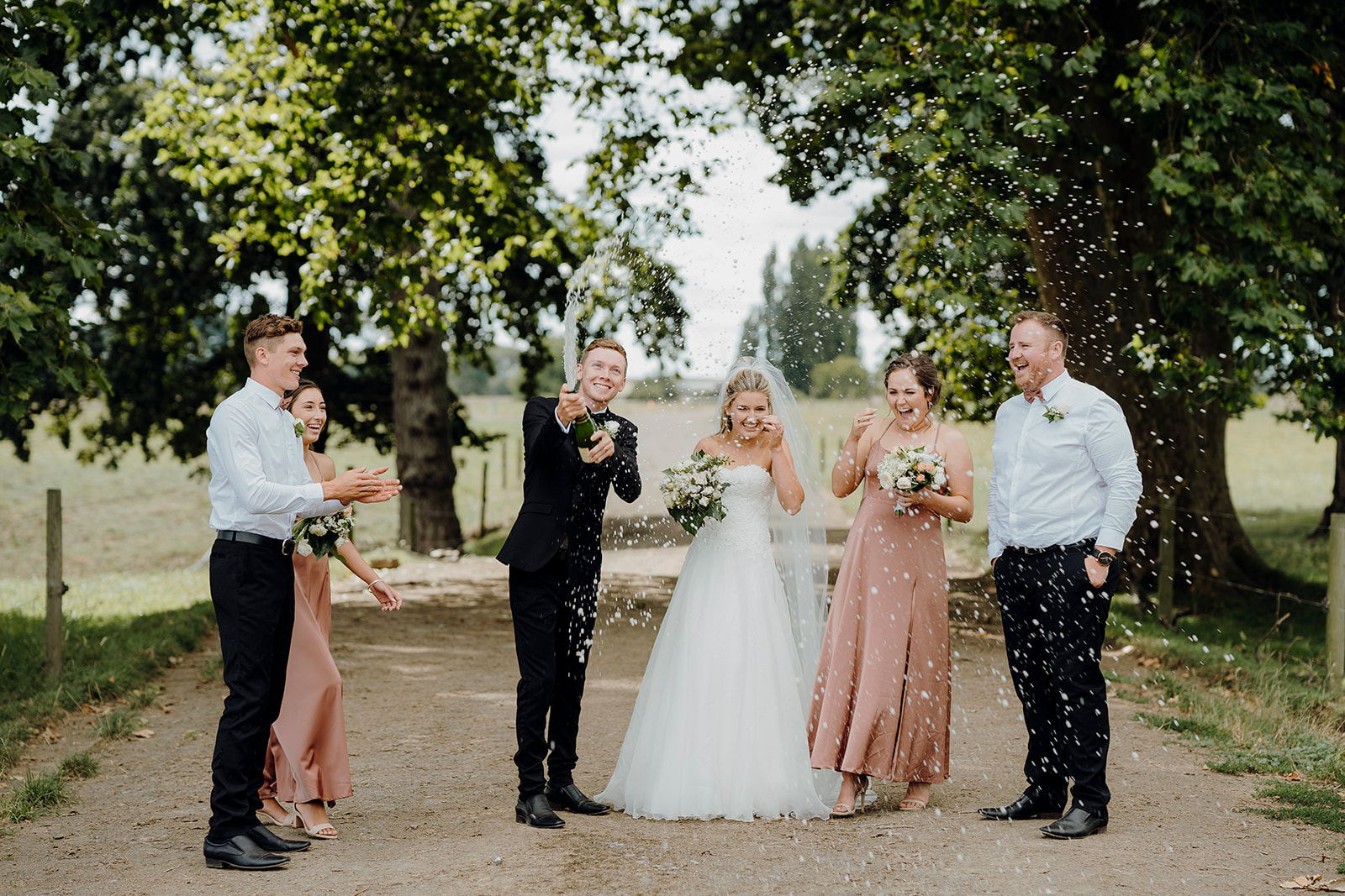 A bride and groom and their wedding party popping a champagne bottle while being photographed by waikato photographer haley adele photography