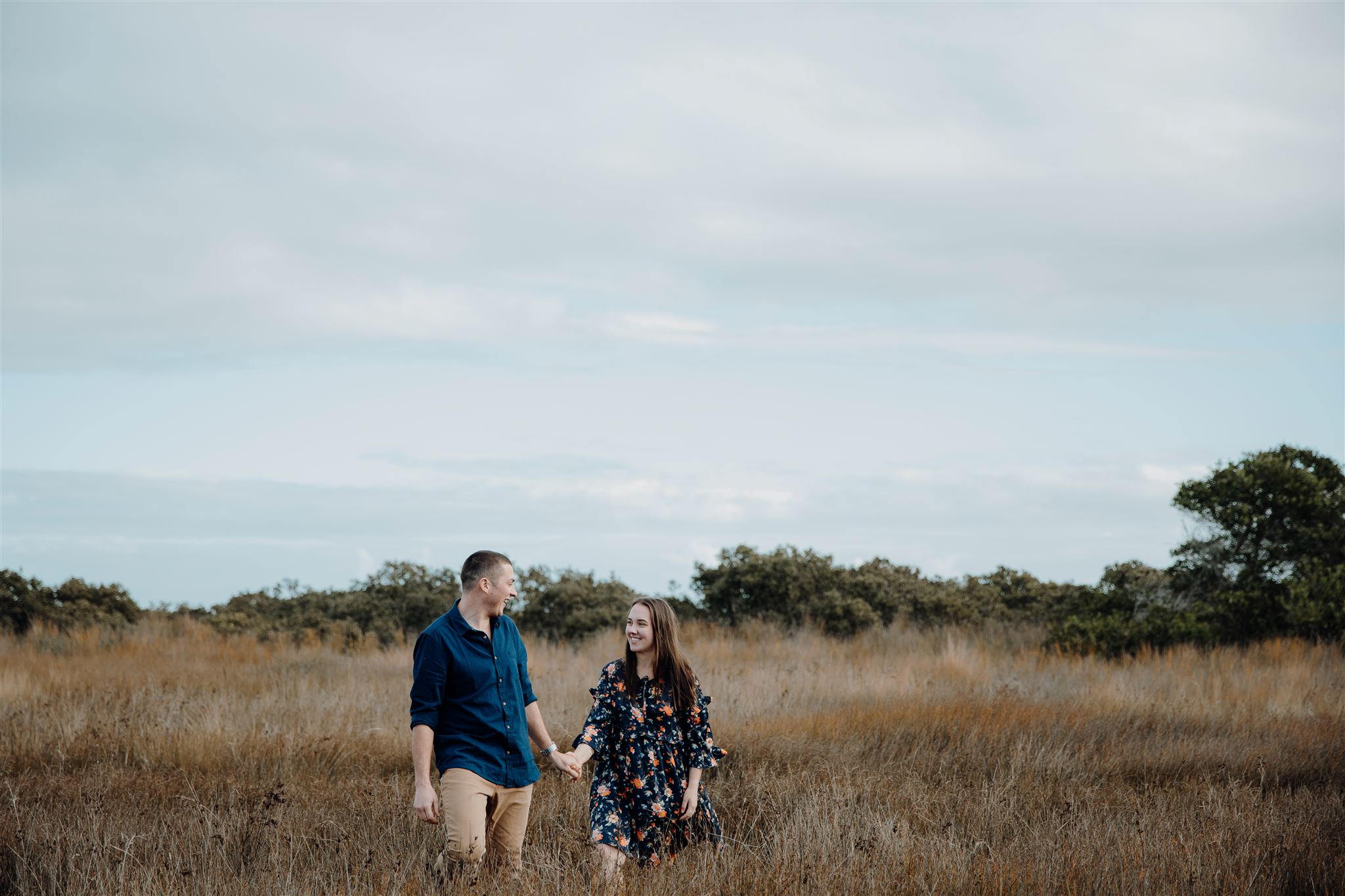 A couple walk through a long field of grass holding hands during an engagement photoshoot by Waikato Photographer Haley Adele Photography