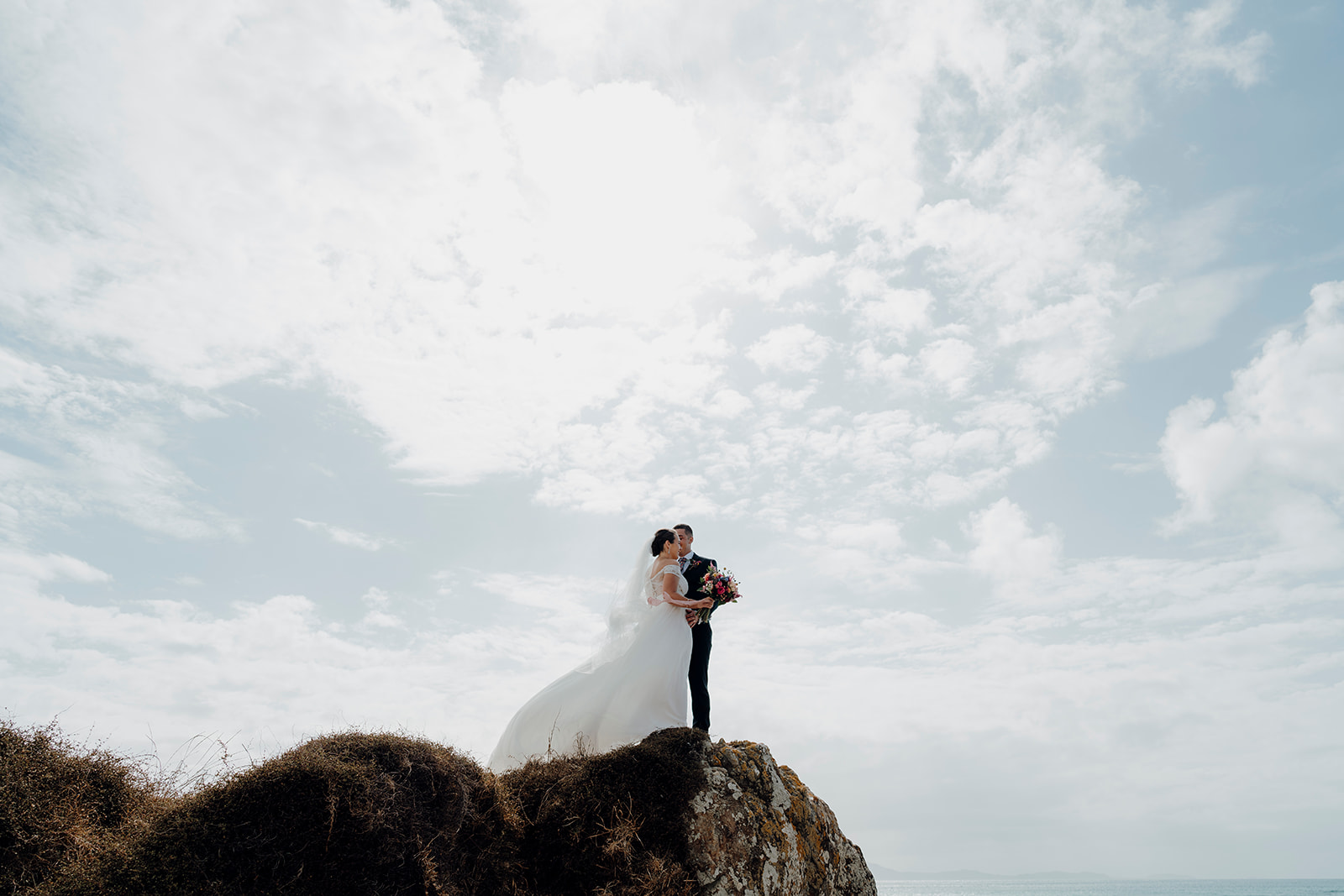 A bride and groom being windswept standing on cliffs during a wedding photoshoot by waikato photographer haley adele photography