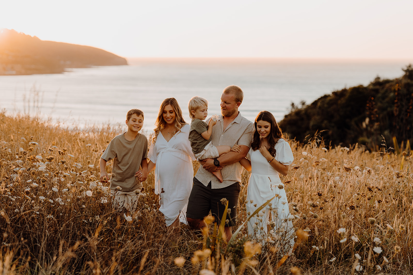 A portrait of a family walking through the long grass in summer during sunset with the Raglan coast in the background photographed by Hamilton Family Photographer Haley Adele Photography