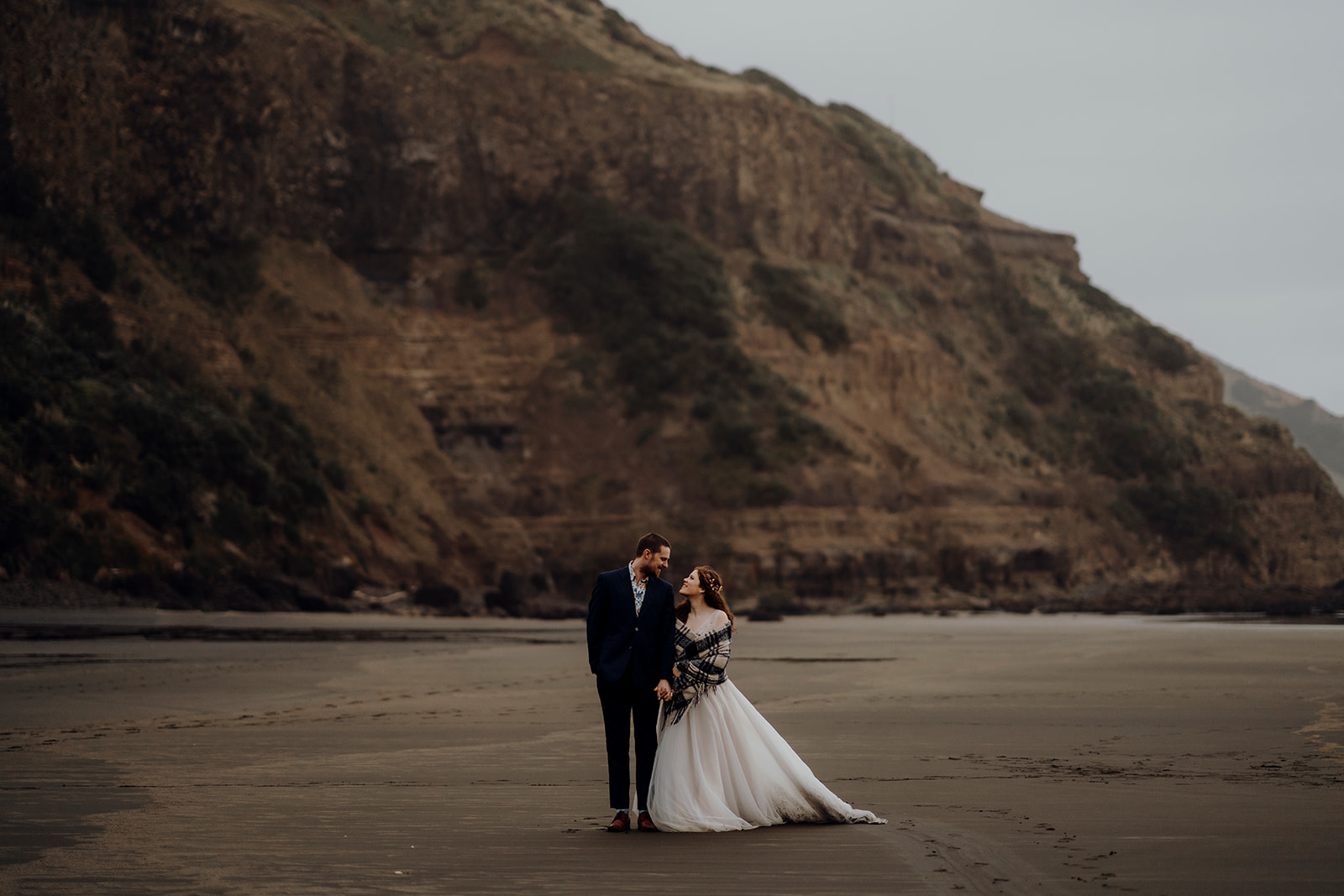 A bride and groom walking along a black sand beach in West Auckland while being photographed by Haley Adele Photography during an elopement style photoshoot