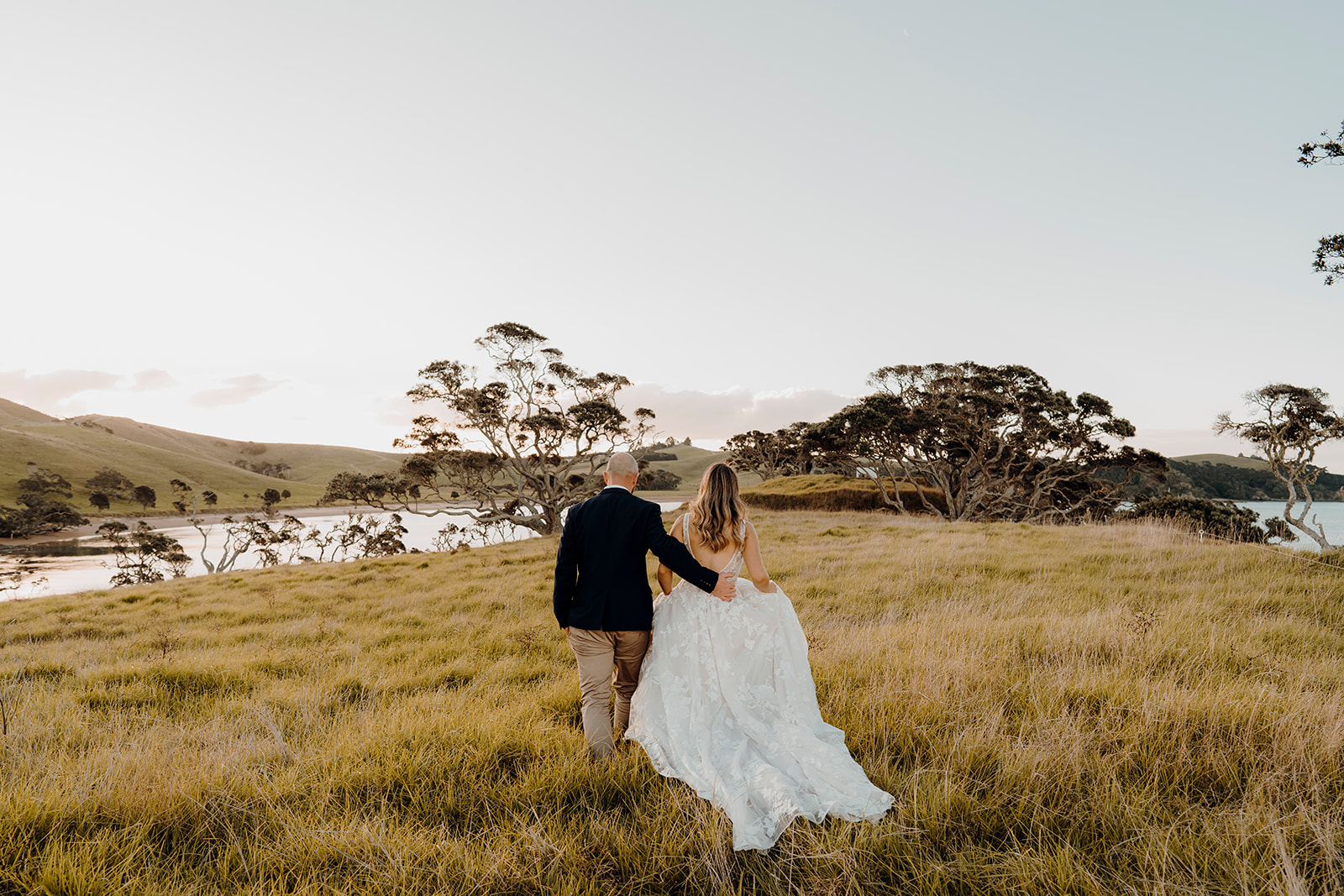A bride and groom walking through the long grass being photographed by Waikato Photographer Haley Adele Photography at Baylys Farm in Northland