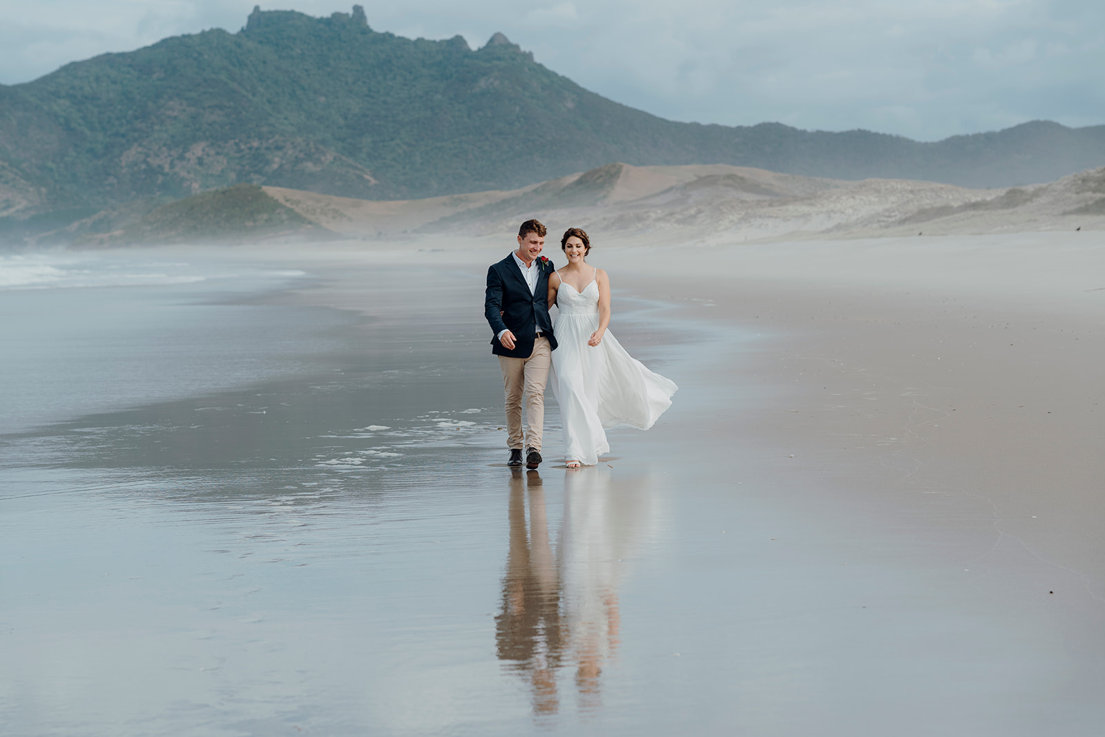 A bride and groom walking along a white sand beach with mountains in the background during their wedding photoshoot with Waikato Wedding Photographer Haley Adele Photography