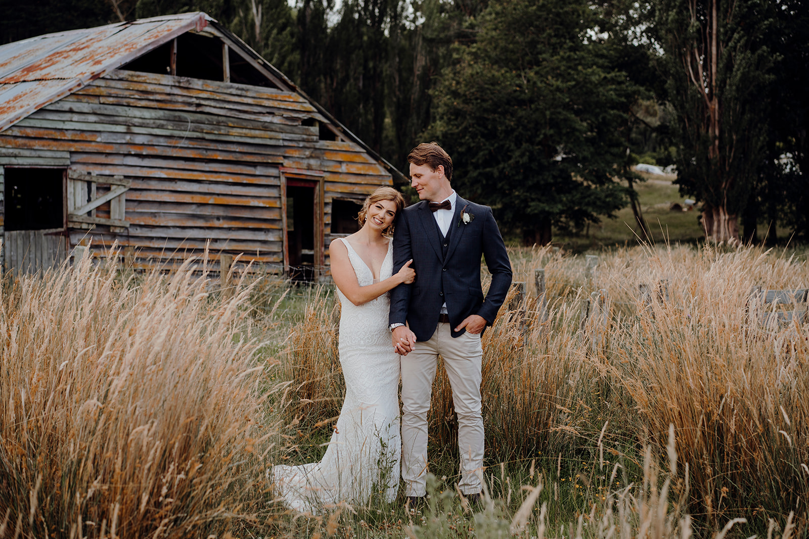 A bride and groom standing in the long grass in front of a rustic shearing shed on a family farm during their wedding photoshoot with waikato photographer haley adele photography