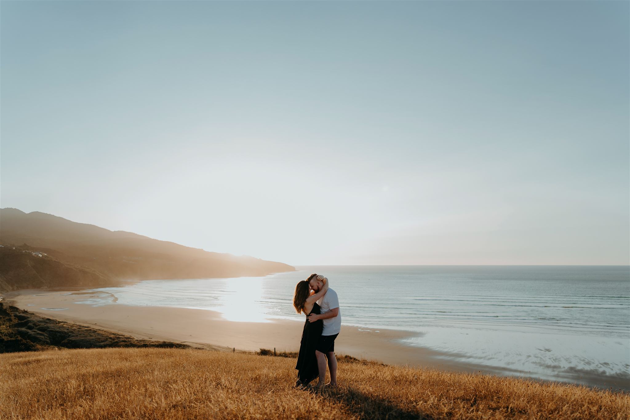 A couple embracing each other on a hill overlooking Raglan beach during a couple photoshoot by Haley Adele Photography.