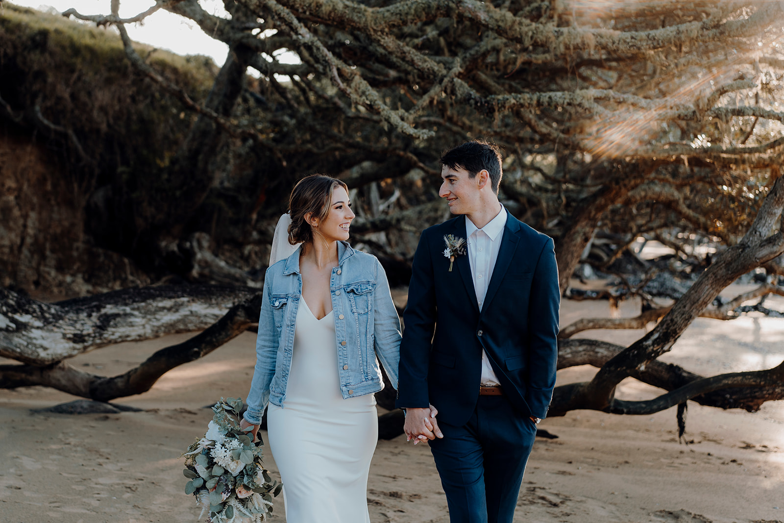 A bride wearing a denim jacket with a groom in a blue suit walking along the private beach at Baylys Farm being photographed by Haley Adele Photography in Northland