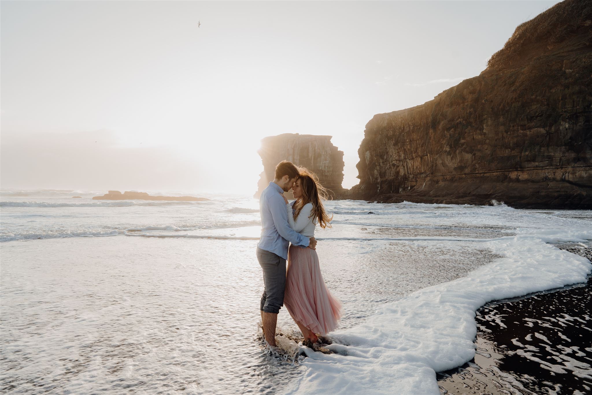 A couple being swept at by the wind at Moukatia Beach in Muriwai during a pre-wedding photoshoot, taken by Haley Adele Photography, a Waikato & Bay of Plenty Wedding Photographer