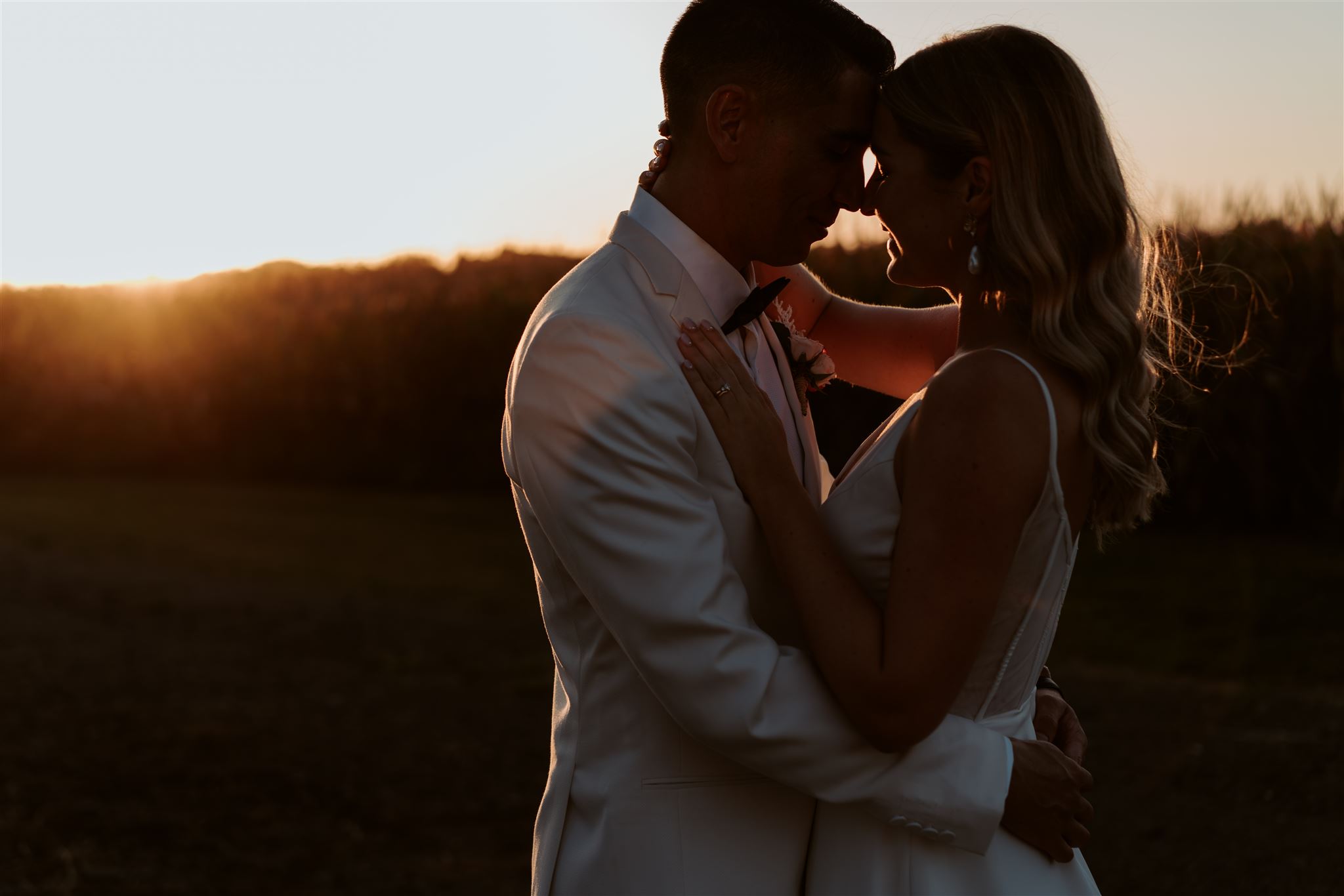 A wedding photograph of a bride and groom from the waist up, showing the sunset over the corn fields at Narrows Landing, a Waikato Wedding Venue, photographed by Haley Adele Photography.