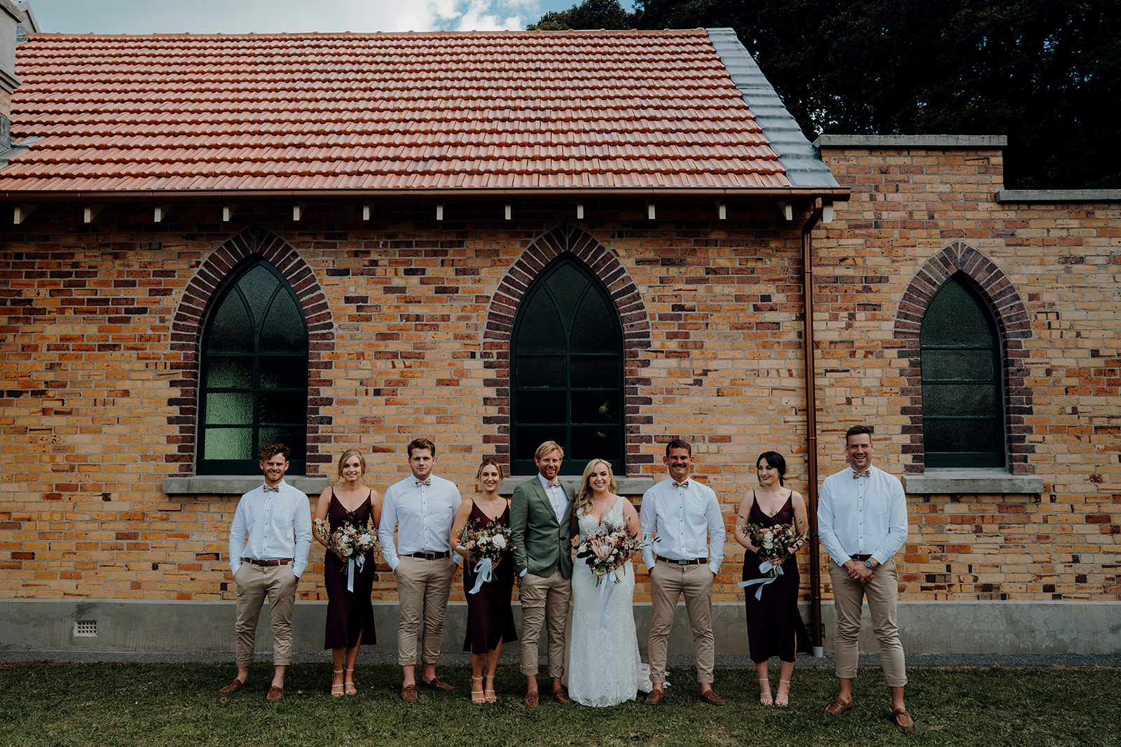 A wedding party with the bride and groom being photographed against the brick walls of the church by Waikato photographer Haley Adele Photography
