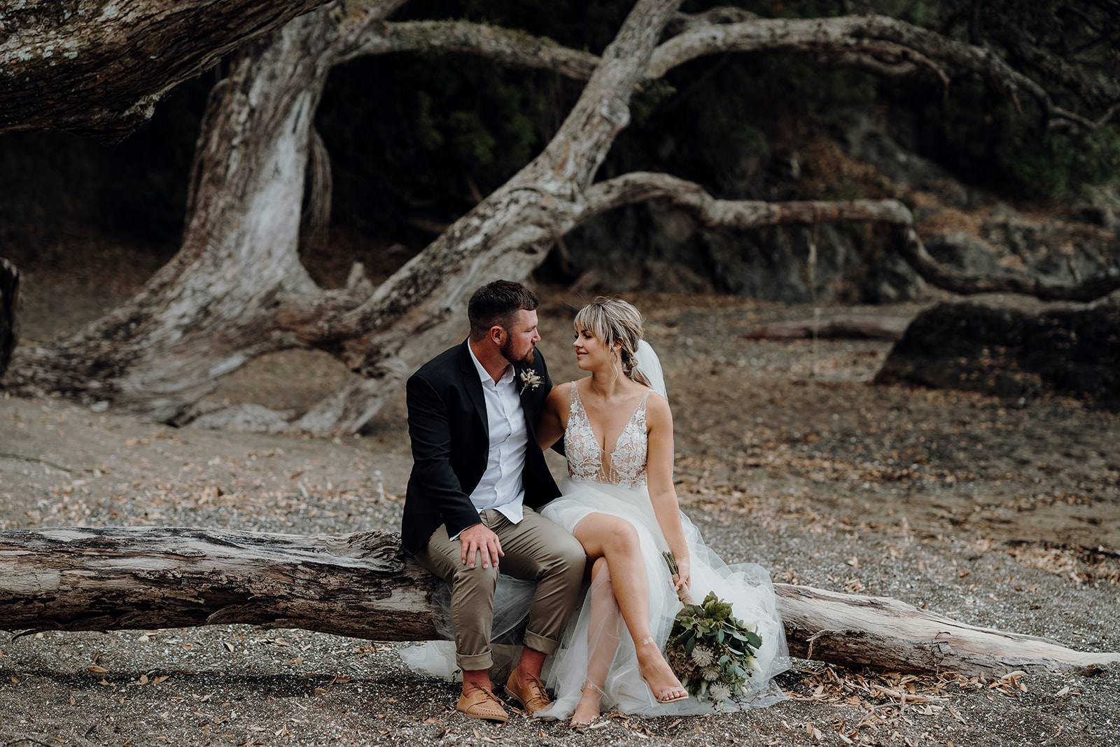 A bride and groom sitting on a log on a beach with dark sand while being photographed by Waikato photographer Haley Adele Photography during their wedding portrait photoshoot