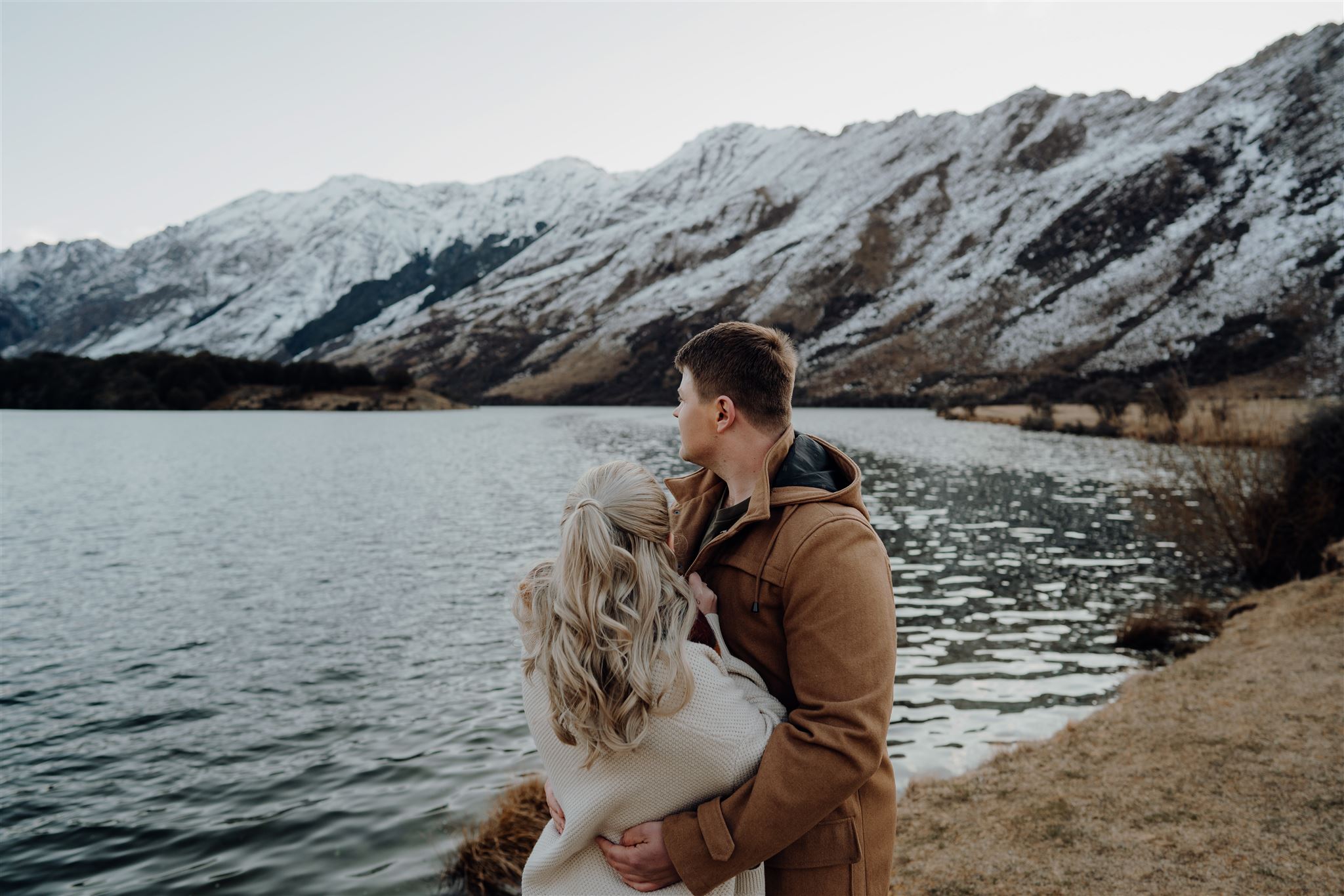 A couple standing in front of Moke Lake in Queenstown with snowy mountains in the background, photographed by Haley Adele Photography