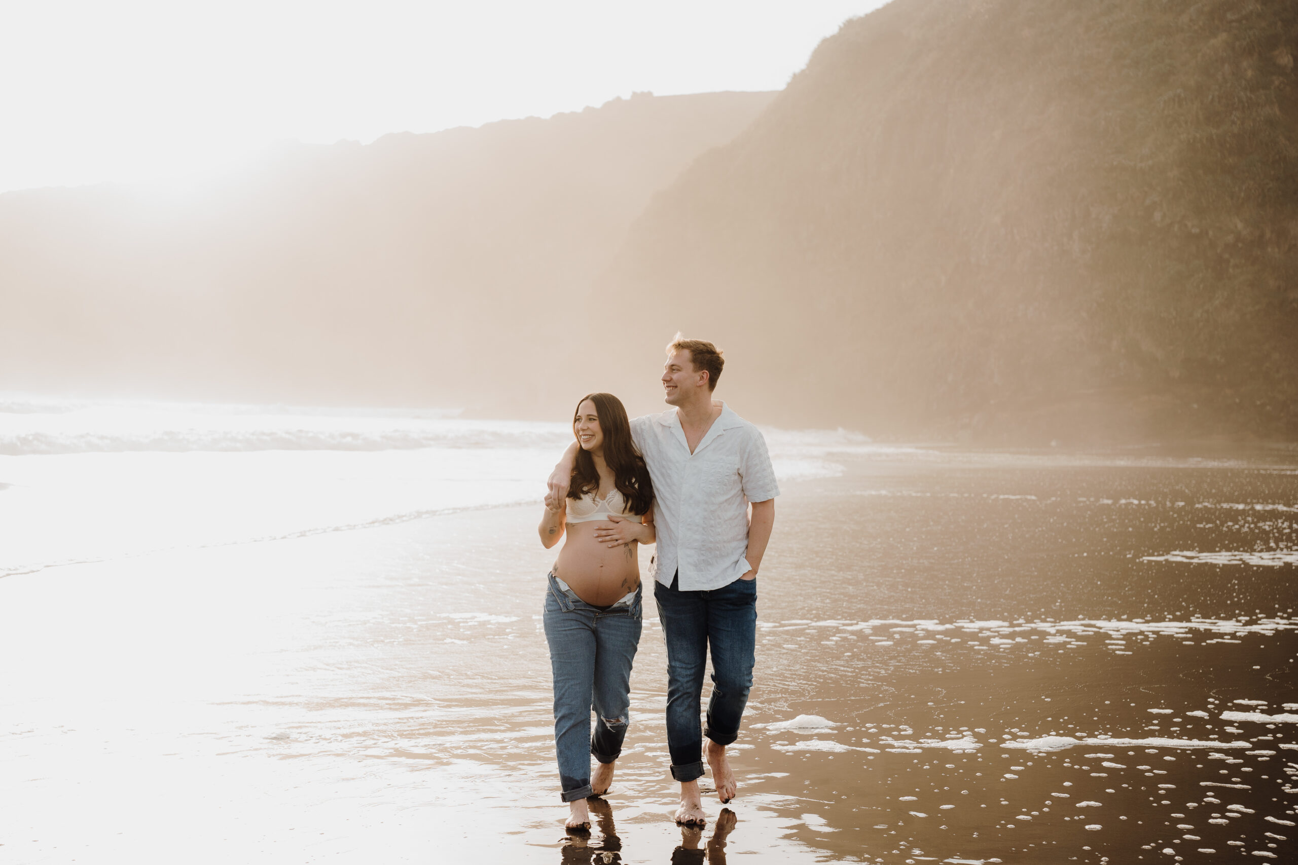 Emma and Josh walking through shallow water at Ruapuke Beach, his arm around her shoulders while she holds her baby bump, dressed in casual denim and lace at sunset.