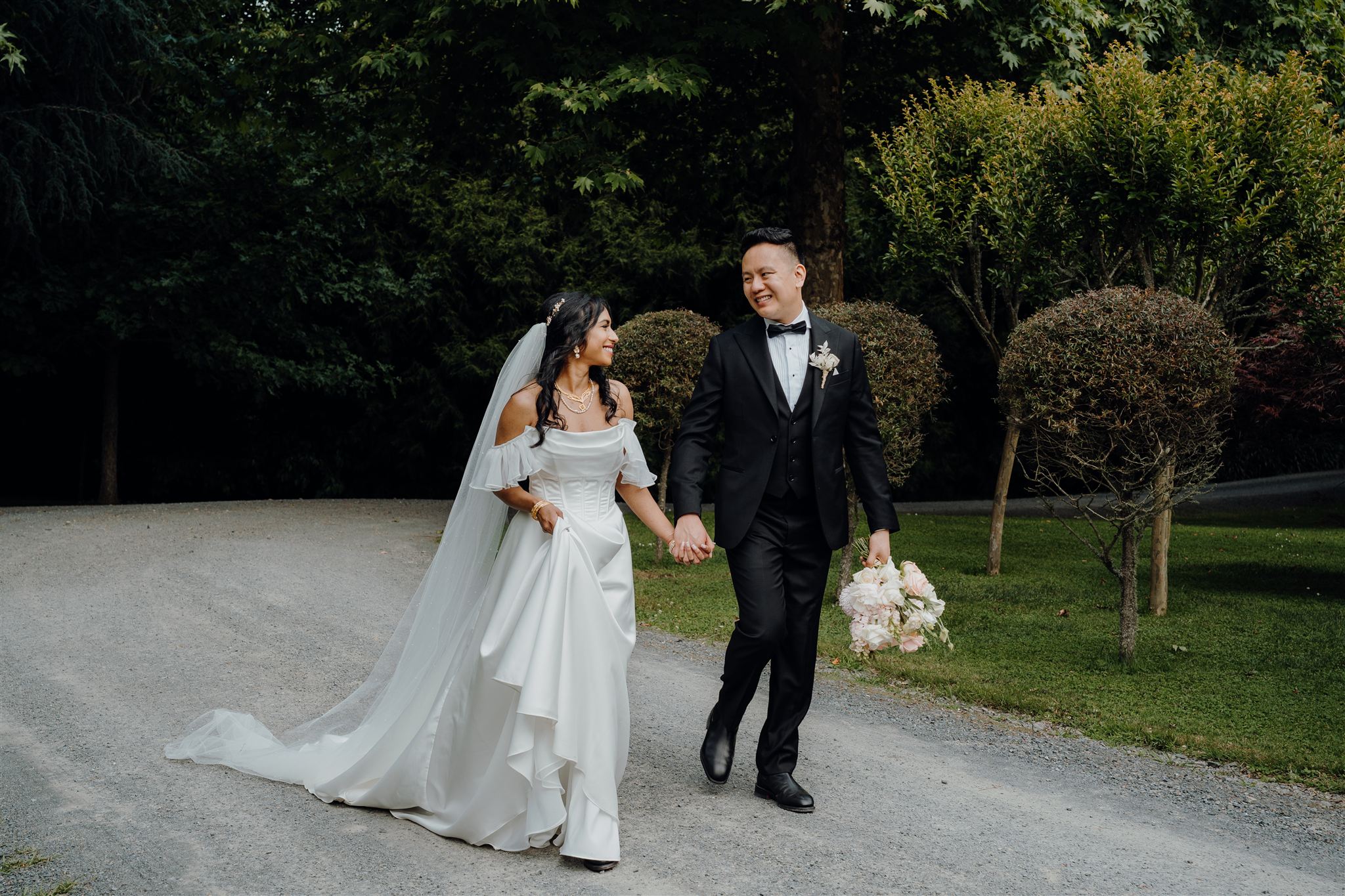 Lakna and Jeffrey walk hand in hand along the tree-lined driveway at The Tree Church and Gardens in Ohaupo. Lakna lifts the front of her dress slightly, the folds of fabric catching the light as they smile at each other.