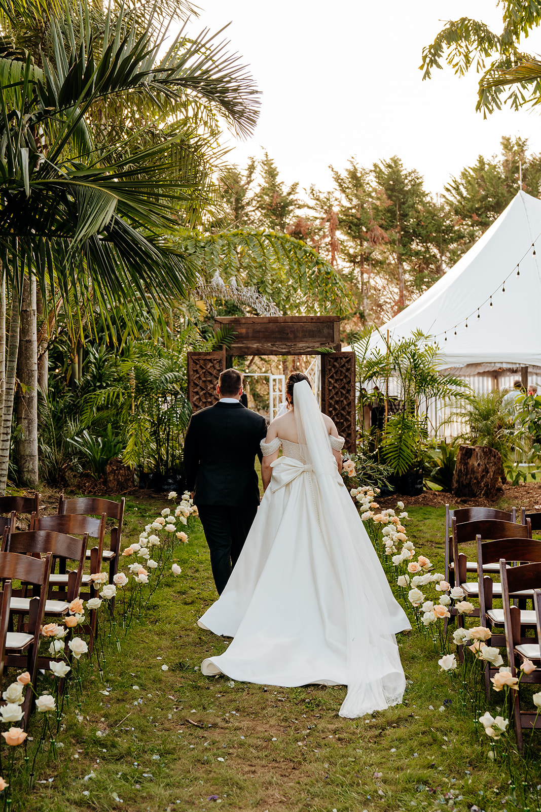 Joshua and India stand together in the aisle of their outdoor wedding, surrounded by pink and white roses on either side. The sunlight filters through tropical ferns behind them, with the marquee softly visible in the distance.