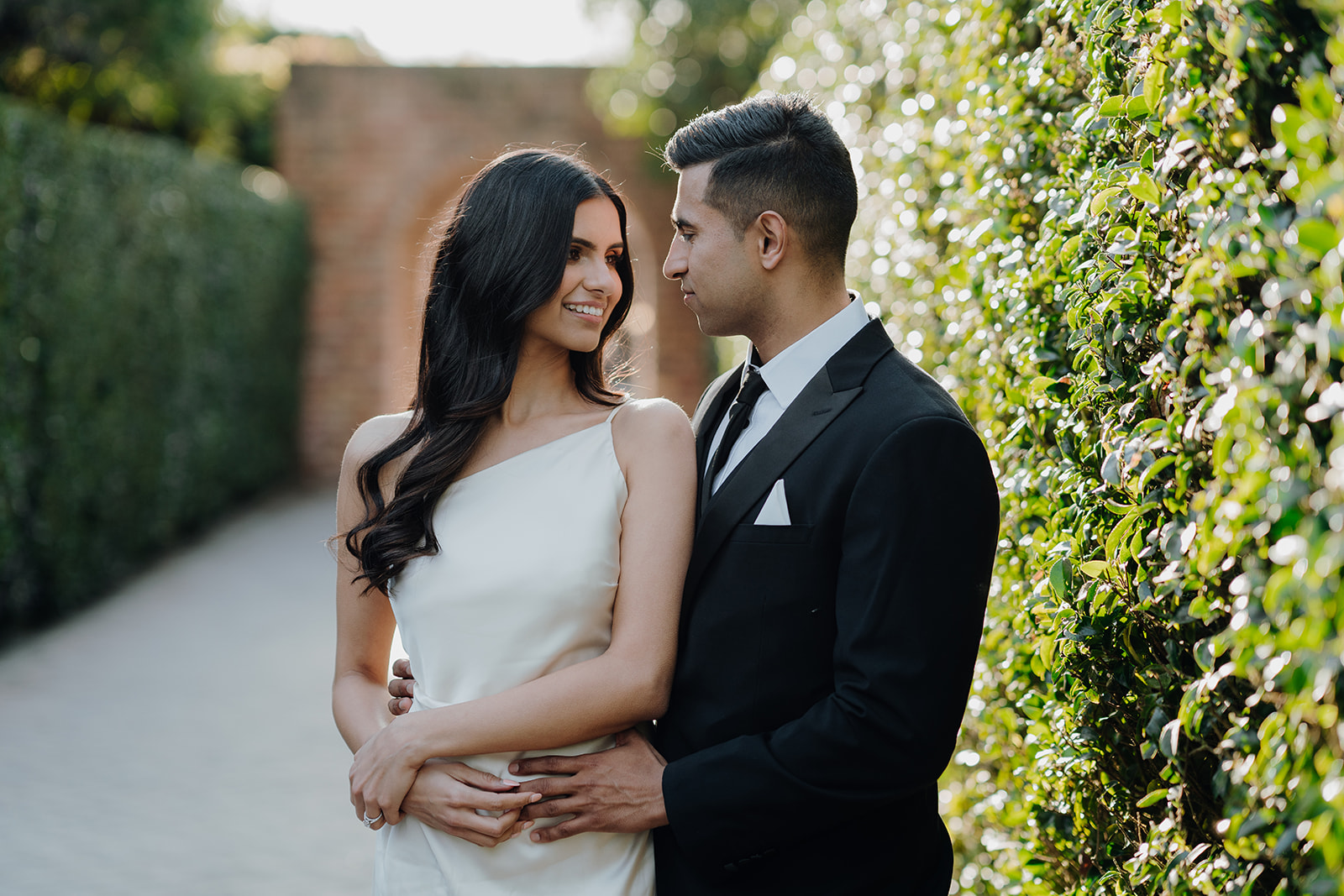 Ritu and Harmon walk hand in hand through the hedge-lined walkway at Hamilton Gardens, dressed in a white dress and suit, surrounded by lush greenery in the enclosed gardens.