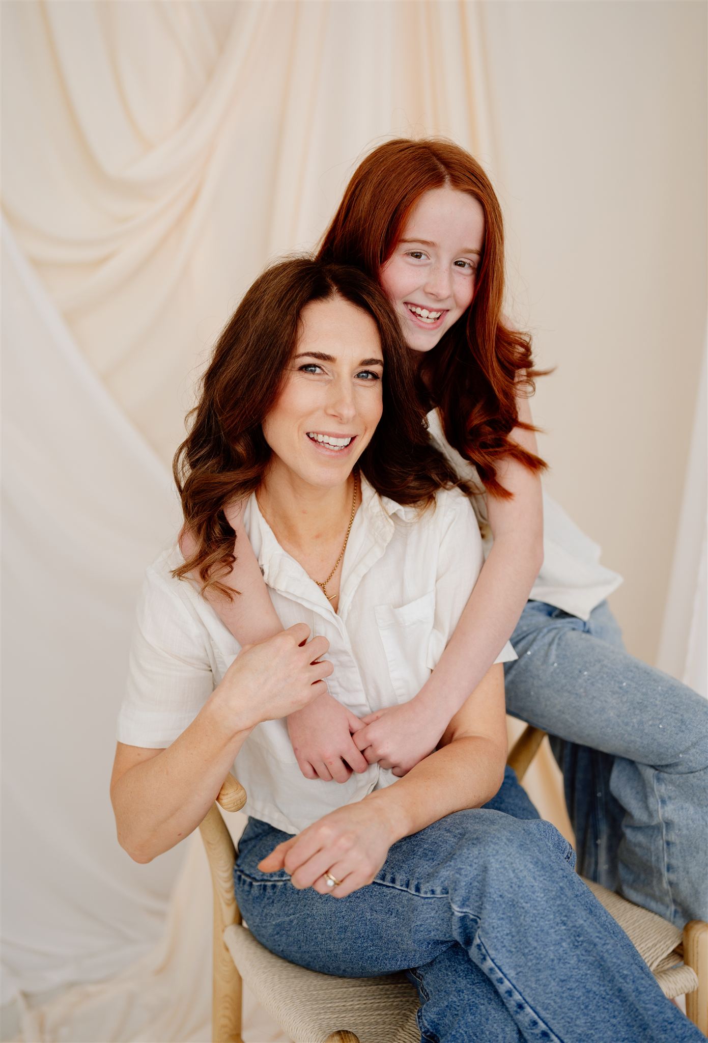 Ruth and her daughter Ruby wear matching jeans and white tops, posing against a draped cream and white fabric backdrop in the studio. Ruby wraps her arms around her mum in a warm, loving hug.